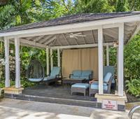 Poolside cabana at Sandalwood House offering a shaded seating area, ceiling fan, and lush garden views