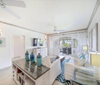 Open and airy living room in Seabreeze Penthouse at Sugar Hill Resort, Barbados, decorated with coastal-inspired decor, white furnishings, and a large television, flowing seamlessly onto the balcony with beautiful garden views.