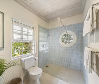 Image of a beautifully designed bathroom at Seascape, Barbados: A charming bathroom with ocean-inspired decor, featuring a glass-enclosed shower with blue tile accents, a round porthole-style window offering lush garden views, and subtle coastal details perfect for a serene holiday rental experience in Barbados.