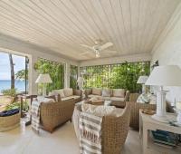 The covered patio lounge at Seascape in Barbados offers comfortable seating, surrounded by lush tropical plants and partial sea views, creating a cozy retreat for holiday villa guests.