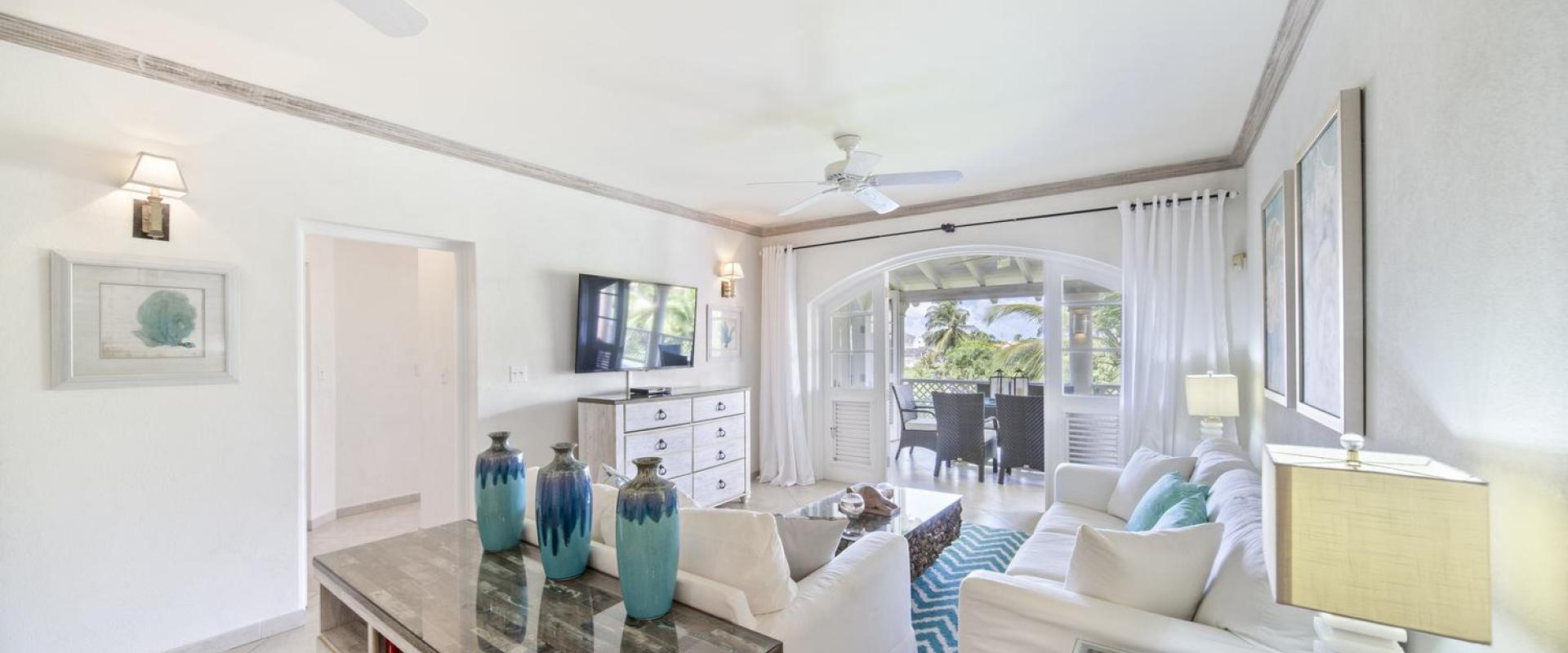 Open and airy living room in Seabreeze Penthouse at Sugar Hill Resort, Barbados, decorated with coastal-inspired decor, white furnishings, and a large television, flowing seamlessly onto the balcony with beautiful garden views.